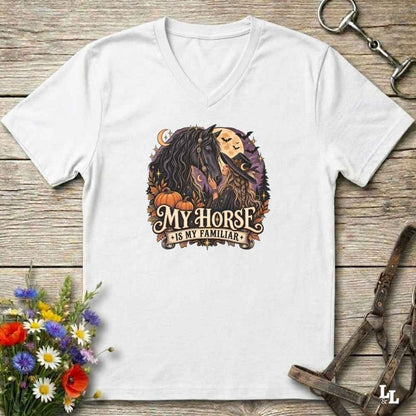 A white V-neck t-shirt laid flat on a rustic wooden surface, featuring a graphic of a horse and a witch with the text 'MY HORSE IS MY FAMILIAR'. The shirt is styled with equestrian accessories (a halter and bit) and autumn-themed flowers.