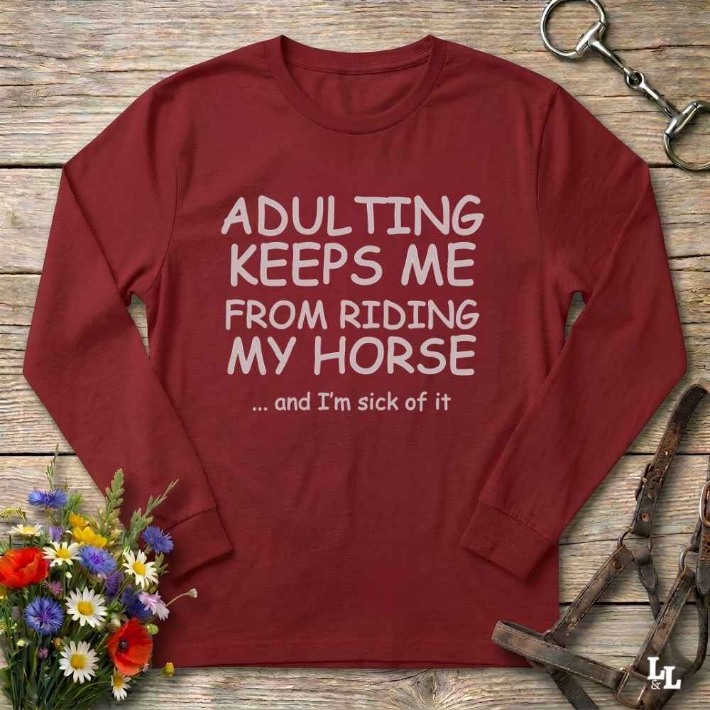 A long-sleeve crewneck t-shirt in maroon red with white text that reads 'ADULTING KEEPS ME FROM RIDING MY HORSE .
