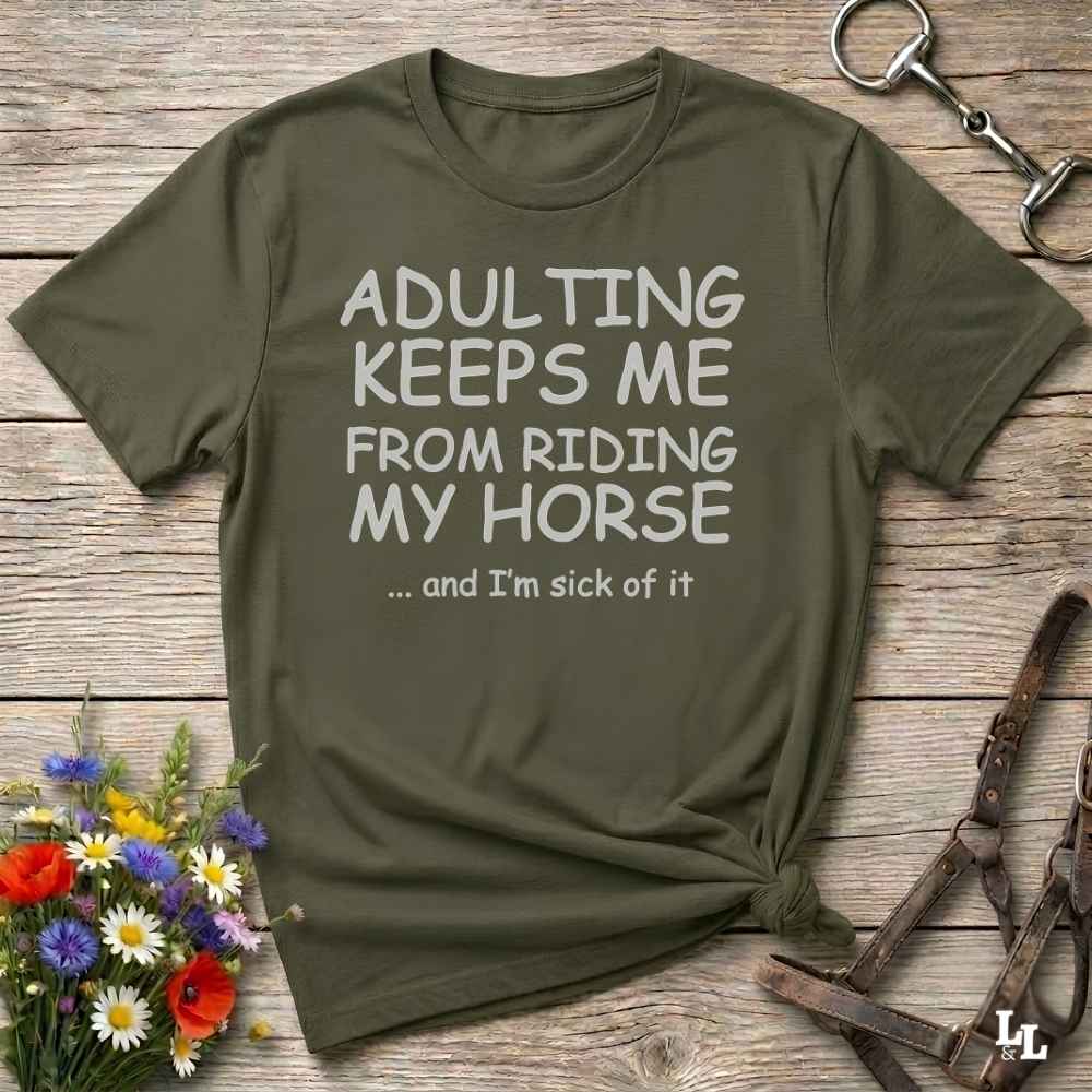 An olive green graphic t-shirt laid flat on a rustic wooden background, styled with equestrian props like a leather bridle and a horse bit, along with small wildflowers. The t-shirt features the text 'ADULTING KEEPS ME FROM RIDING MY HORSE .