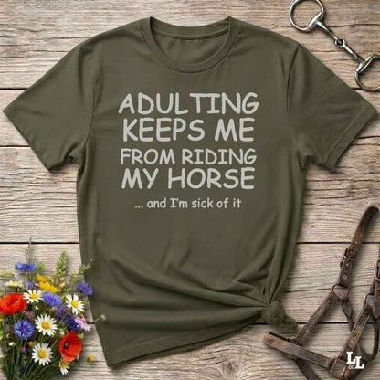 An olive green graphic t-shirt laid flat on a rustic wooden background, styled with equestrian props like a leather bridle and a horse bit, along with small wildflowers. The t-shirt features the text 'ADULTING KEEPS ME FROM RIDING MY HORSE .