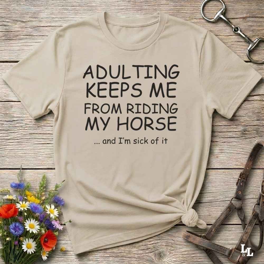 A beige graphic t-shirt with the text 'ADULTING KEEPS ME FROM RIDING MY HORSE .
