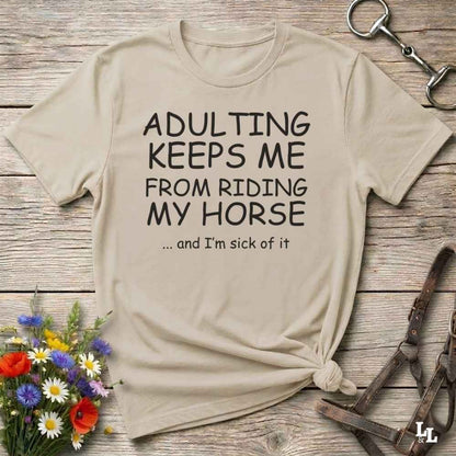 A beige graphic t-shirt with the text 'ADULTING KEEPS ME FROM RIDING MY HORSE .