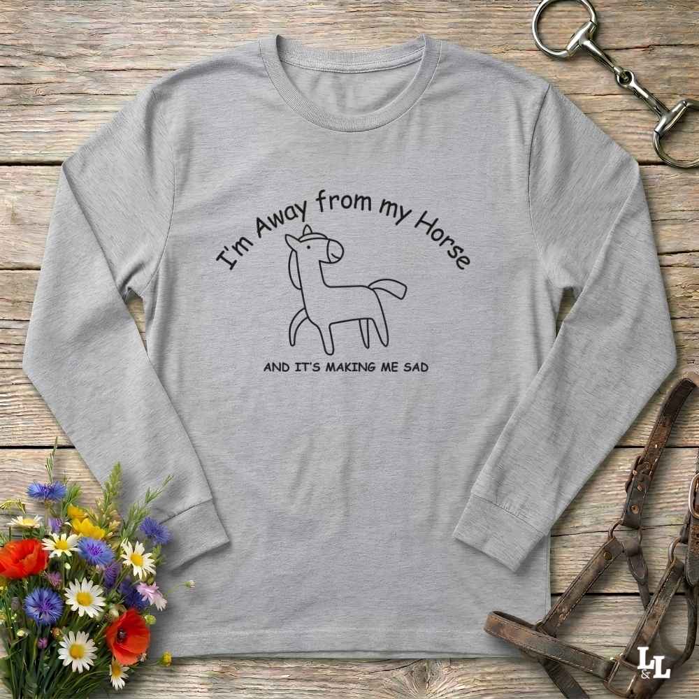 A light gray long-sleeve t-shirt with a black graphic of a sad horse and the text 'I'm Away from my Horse AND IT'S MAKING ME SAD', displayed on a rustic wooden background with equestrian accessories and flowers.