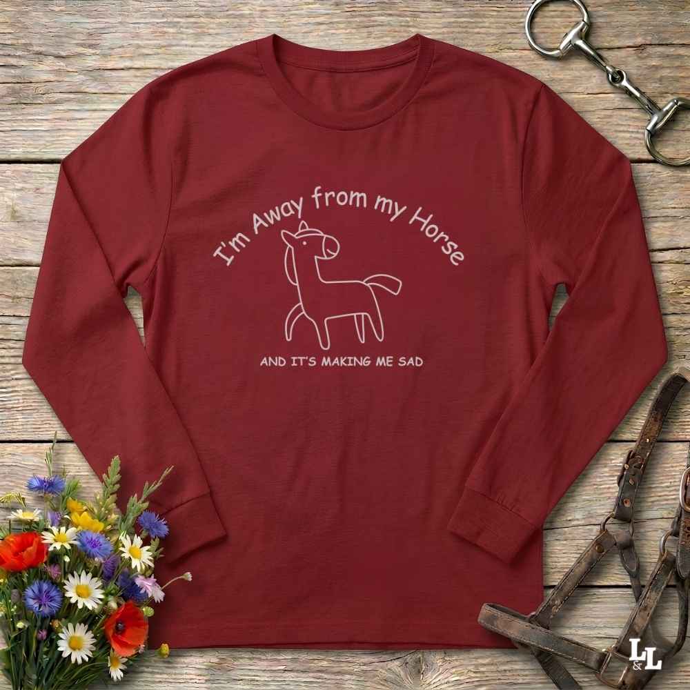 A long-sleeve crewneck t-shirt in maroon red, laid flat on a rustic wooden surface. The shirt features a white line art graphic of a horse with the text 'I'm Away from my Horse AND IT'S MAKING ME SAD'.