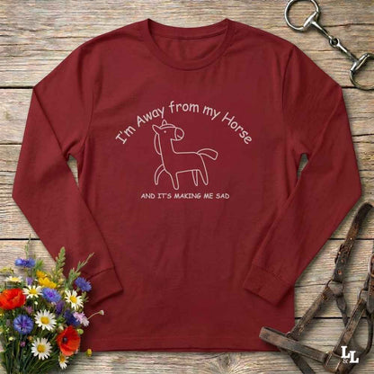 A long-sleeve crewneck t-shirt in maroon red, laid flat on a rustic wooden surface. The shirt features a white line art graphic of a horse with the text 'I'm Away from my Horse AND IT'S MAKING ME SAD'.