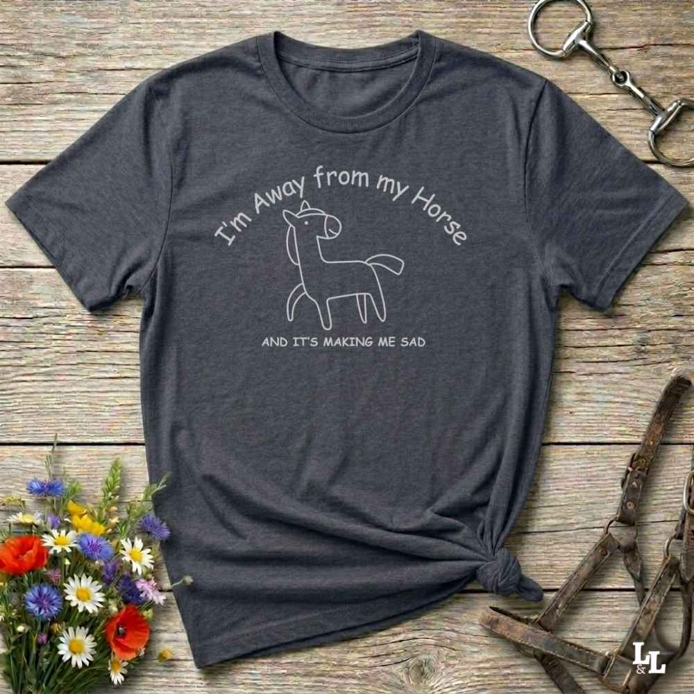 A dark heather gray graphic t-shirt laid flat on a rustic wooden surface, styled with a small bouquet of wildflowers, a horse halter, and a metal horse tack in the corners. The t-shirt features a white line art graphic of a sad horse with the text 'I'm Away from my Horse AND IT'S MAKING ME SAD'.