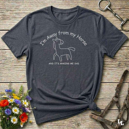 A dark heather gray graphic t-shirt laid flat on a rustic wooden surface, styled with a small bouquet of wildflowers, a horse halter, and a metal horse tack in the corners. The t-shirt features a white line art graphic of a sad horse with the text 'I'm Away from my Horse AND IT'S MAKING ME SAD'.