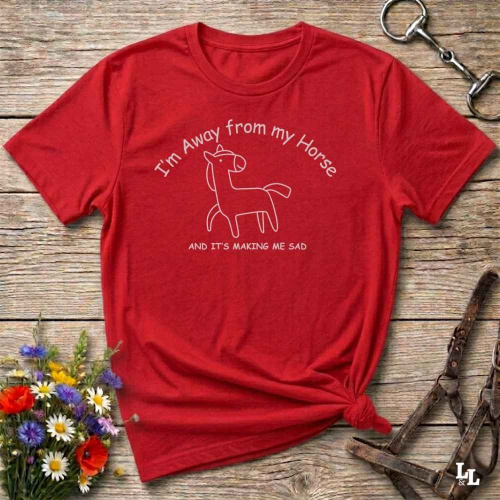 A red short-sleeve graphic T-shirt laid flat on a rustic wooden surface, with a white line drawing of a horse and the text 'I'm Away from my Horse AND IT'S MAKING ME SAD.' Decorative flowers and equestrian hardware are placed around the shirt.