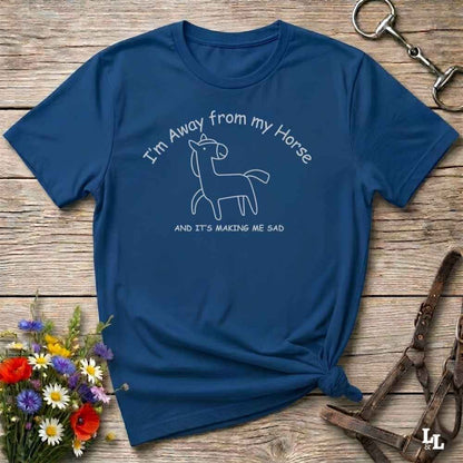 A navy blue short-sleeve graphic T-shirt laid flat on a rustic wooden surface, with white text reading 'I'm Away from my Horse AND IT'S MAKING ME SAD' and a simple line drawing of a horse. The shirt is styled with flowers, a horse bit, and a leather strap around the scene.