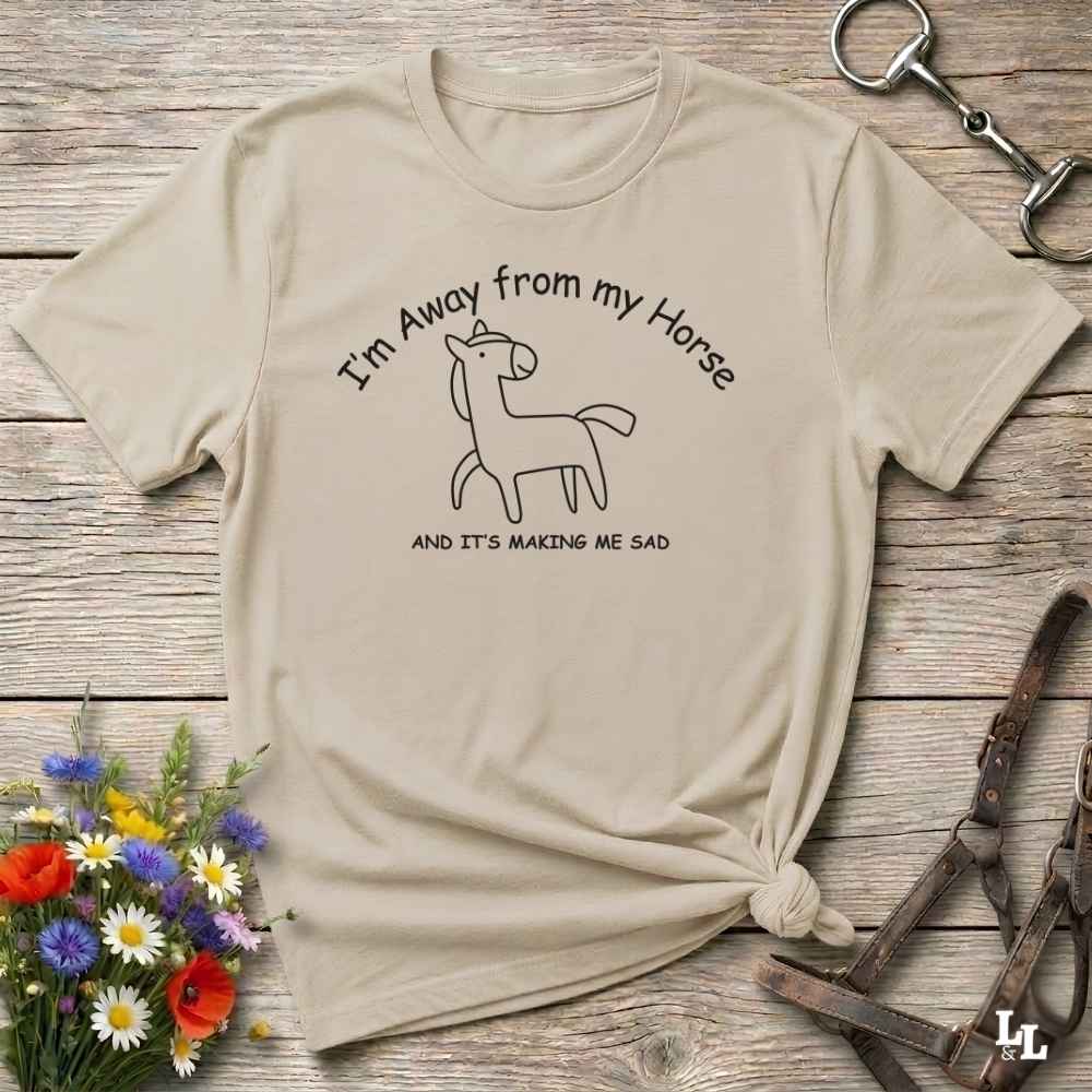 A beige graphic t-shirt with a black line art horse and the text 'I'm Away from my Horse AND IT'S MAKING ME SAD', styled in a flat-lay on a rustic wooden background with flowers and horse tack.