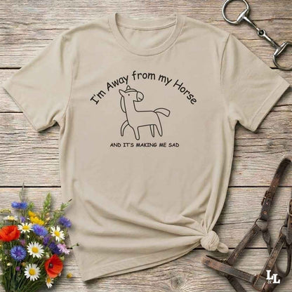 A beige graphic t-shirt with a black line art horse and the text 'I'm Away from my Horse AND IT'S MAKING ME SAD', styled in a flat-lay on a rustic wooden background with flowers and horse tack.