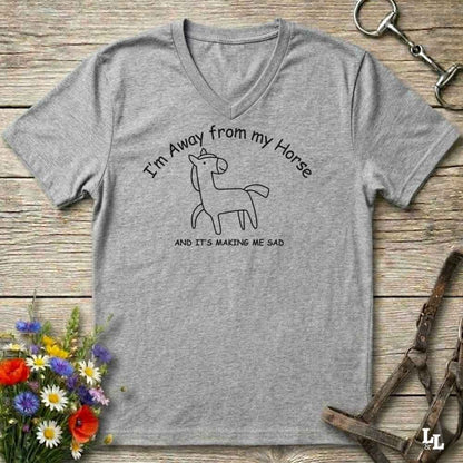 A heather gray V-neck t-shirt with a black line art graphic of a horse and the text 'I'm Away from my Horse AND IT'S MAKING ME SAD', displayed on a rustic wooden background with equestrian-themed props.