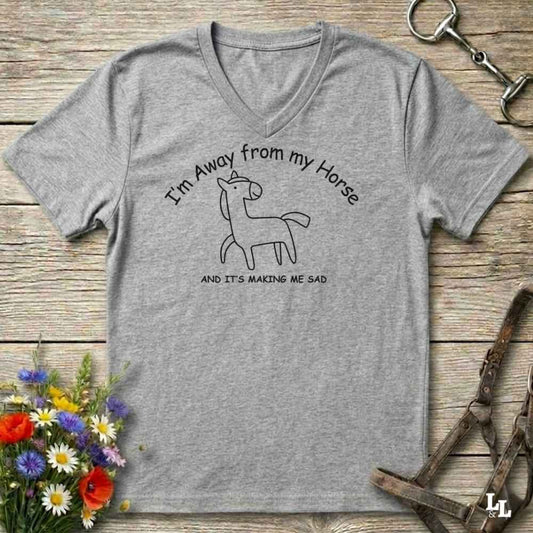 A heather gray V-neck t-shirt with a black line art graphic of a horse and the text 'I'm Away from my Horse AND IT'S MAKING ME SAD', displayed on a rustic wooden background with equestrian-themed props.