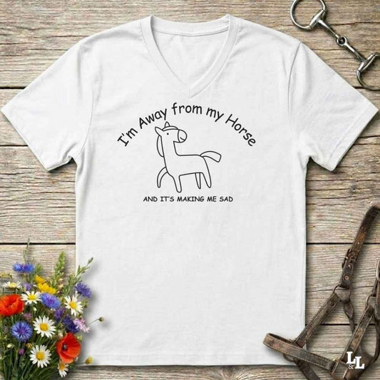 A white  V-neck t-shirt with a black line art graphic of a horse and the text 'I'm Away from my Horse AND IT'S MAKING ME SAD', displayed on a rustic wooden background with equestrian-themed props.