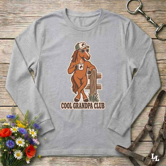 A heather gray long-sleeve t-shirt with a graphic of a cartoon horse wearing a cowboy hat and sunglasses, holding a mug, with the text 'COOL GRANDPA CLUB' underneath. The shirt is displayed in a flat-lay arrangement on a rustic wooden background with decorative equestrian hardware and a flower bouquet.