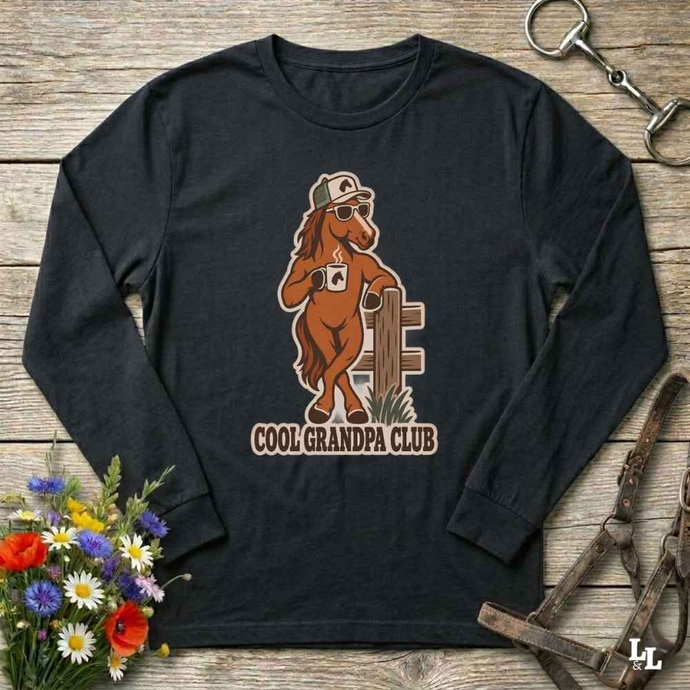 A black long-sleeve t-shirt with a cartoon graphic of a brown horse wearing sunglasses and a cap, next to a fence, with the text 'COOL GRANDPA CLUB' printed below. The shirt is displayed flat on a rustic wooden background with equestrian accessories and flowers.