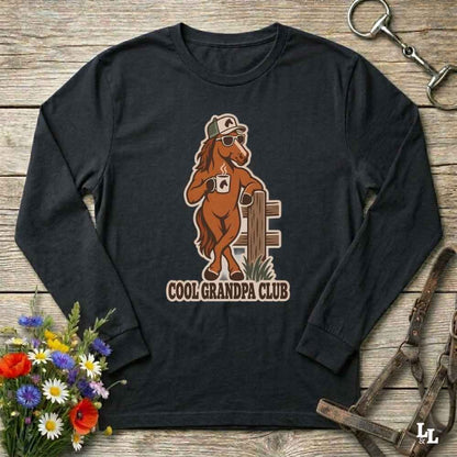 A black long-sleeve t-shirt with a cartoon graphic of a brown horse wearing sunglasses and a cap, next to a fence, with the text 'COOL GRANDPA CLUB' printed below. The shirt is displayed flat on a rustic wooden background with equestrian accessories and flowers.