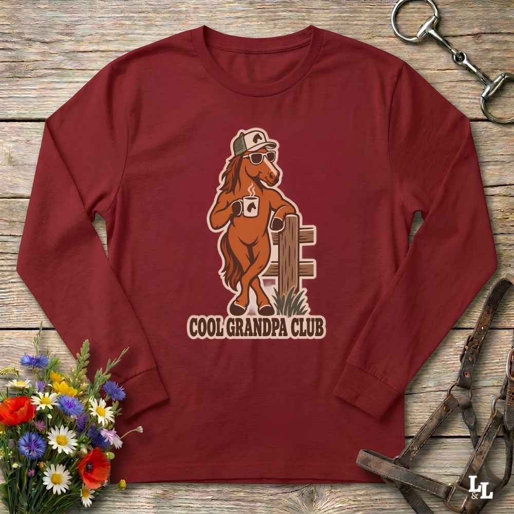 A long-sleeve crewneck shirt in maroon red, laid flat on a rustic wooden surface. The shirt features a graphic of a cartoon brown horse wearing a white cap and sunglasses, holding a mug next to a wooden fence, with the text 'COOL GRANDPA CLUB' underneath.