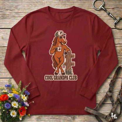 A long-sleeve crewneck shirt in maroon red, laid flat on a rustic wooden surface. The shirt features a graphic of a cartoon brown horse wearing a white cap and sunglasses, holding a mug next to a wooden fence, with the text 'COOL GRANDPA CLUB' underneath.