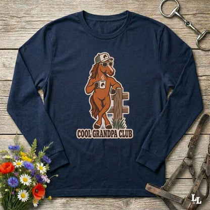 A navy blue long-sleeve t-shirt with a vintage-style graphic of a cool brown horse wearing sunglasses and a cap, holding a coffee mug next to a fence, with the text 'COOL GRANDPA CLUB' printed below. The shirt is displayed flat on a rustic wooden background with equestrian and floral props.