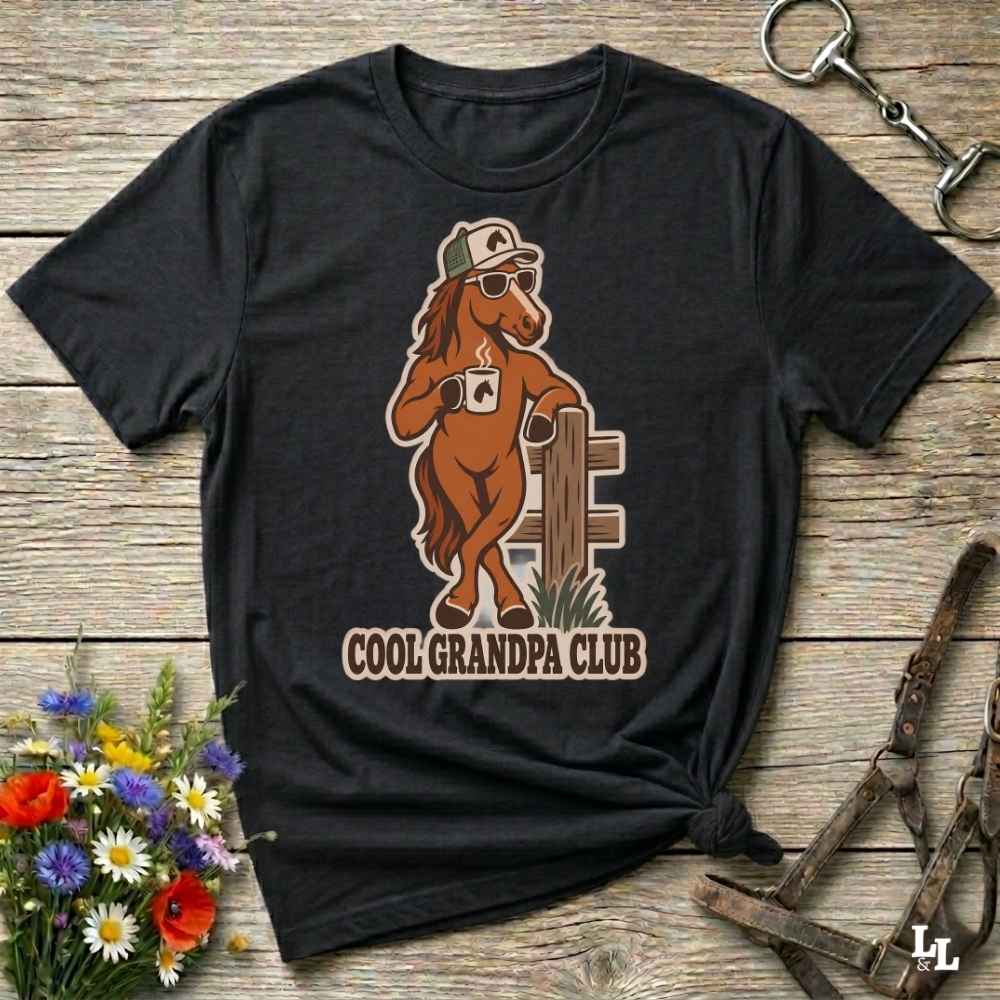 A black short-sleeve graphic T-shirt laid flat on a rustic wooden surface, featuring a brown cartoon horse wearing a cap and sunglasses with the text 'COOL GRANDPA CLUB.' Decorative flowers and horse tack are placed around the shirt.