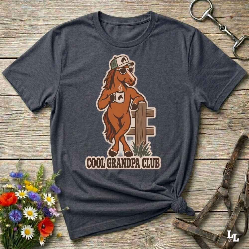 A dark heather gray graphic t-shirt laid flat on a rustic wooden surface, styled with a small bouquet of flowers, a horse bit, and horse tack. The t-shirt features a graphic of a cool brown horse wearing sunglasses, a cap, and a mug with the text 'COOL GRANDPA CLUB'.