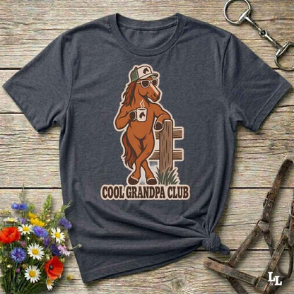 A dark heather gray graphic t-shirt laid flat on a rustic wooden surface, styled with a small bouquet of flowers, a horse bit, and horse tack. The t-shirt features a graphic of a cool brown horse wearing sunglasses, a cap, and a mug with the text 'COOL GRANDPA CLUB'.
