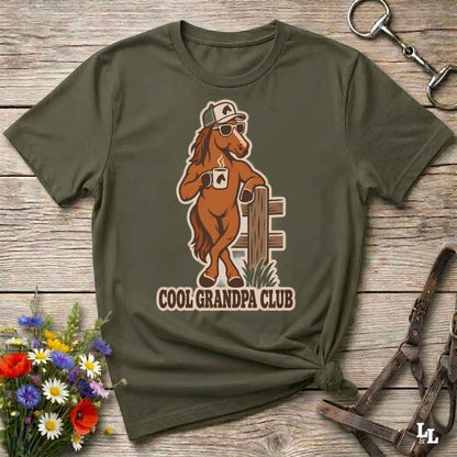 An olive green graphic t-shirt laid flat on a rustic wooden surface, styled with a small bouquet of flowers, a horse bit, and a leather bridle. The t-shirt features a cartoon graphic of a brown horse wearing sunglasses and a cap, leaning on a fence, with the text 'COOL GRANDPA CLUB' underneath.