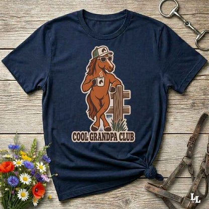 A navy blue graphic t-shirt laid flat on a rustic wooden surface, styled with equestrian accessories and a small bouquet of wildflowers. The shirt features a cartoon graphic of a brown horse wearing sunglasses and a cap, leaning on a fence, with the text 'COOL GRANDPA CLUB' underneath.