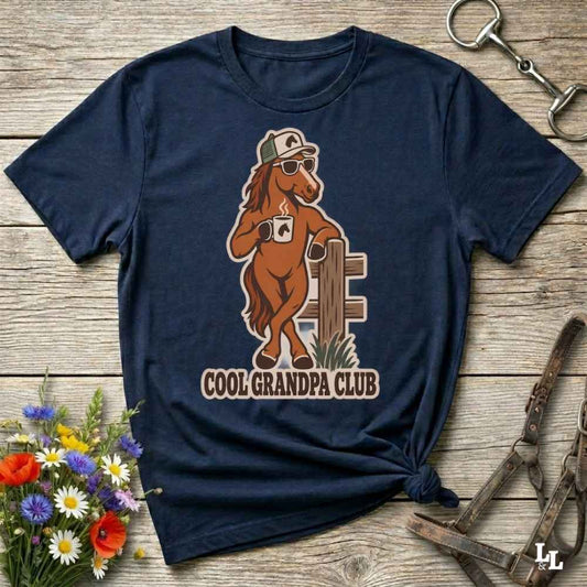 A navy blue graphic t-shirt laid flat on a rustic wooden surface, styled with equestrian accessories and a small bouquet of wildflowers. The shirt features a cartoon graphic of a brown horse wearing sunglasses and a cap, leaning on a fence, with the text 'COOL GRANDPA CLUB' underneath.