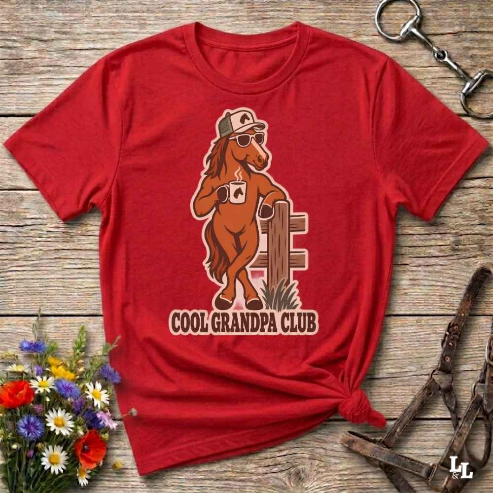 A red short-sleeve graphic T-shirt laid flat on a rustic wooden surface, styled with flowers, tack, and a horse halter. The shirt features a cartoon horse wearing sunglasses and a cap, with the text 'COOL GRANDPA CLUB' below.