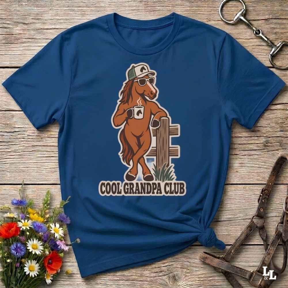 A navy blue graphic t-shirt laid flat on a rustic wooden surface, featuring a cartoon brown horse wearing sunglasses and a cap with the text 'COOL GRANDPA CLUB'. The shirt is styled with equestrian accessories (a halter and a horse bit) and a small bouquet of wildflowers.