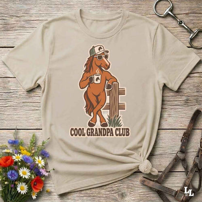 A beige graphic t-shirt laid flat on a rustic wooden surface, styled with equestrian accessories and a small bouquet of wildflowers. The shirt features a cartoon illustration of a brown horse wearing sunglasses and a cap, leaning on a fence, with the text 'COOL GRANDPA CLUB' printed below.