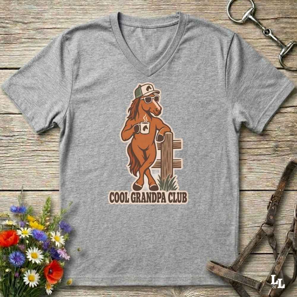A heather gray V-neck t-shirt laid flat on a rustic wooden surface, featuring a cartoon graphic of a cool horse wearing sunglasses and a cap, with the text 'COOL GRANDPA CLUB'. The shirt is styled with equestrian accessories (a horse bit in the top right and horse reins in the bottom right) and a small bouquet of colorful wildflowers in the bottom left.