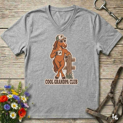 A heather gray V-neck t-shirt laid flat on a rustic wooden surface, featuring a cartoon graphic of a cool horse wearing sunglasses and a cap, with the text 'COOL GRANDPA CLUB'. The shirt is styled with equestrian accessories (a horse bit in the top right and horse reins in the bottom right) and a small bouquet of colorful wildflowers in the bottom left.