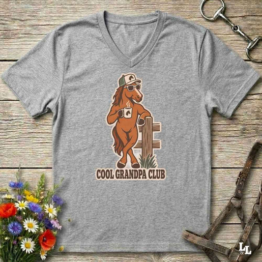 A heather gray V-neck t-shirt laid flat on a rustic wooden surface, featuring a cartoon graphic of a cool horse wearing sunglasses and a cap, with the text 'COOL GRANDPA CLUB'. The shirt is styled with equestrian accessories (a horse bit in the top right and horse reins in the bottom right) and a small bouquet of colorful wildflowers in the bottom left.