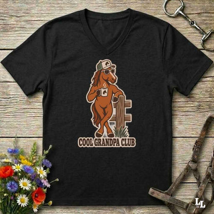 A black V-neck t-shirt laid flat on a rustic wooden surface, featuring a graphic of a brown horse wearing sunglasses and a hat, holding a coffee mug with the text 'COOL GRANDPA CLUB'. The shirt is styled with equestrian accessories (reins and a bit) and a small bouquet of wildflowers.