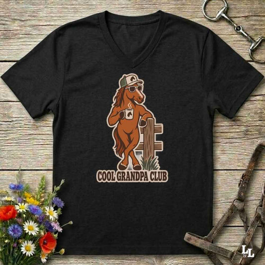 A black V-neck t-shirt laid flat on a rustic wooden surface, featuring a graphic of a brown horse wearing sunglasses and a hat, holding a coffee mug with the text 'COOL GRANDPA CLUB'. The shirt is styled with equestrian accessories (reins and a bit) and a small bouquet of wildflowers.