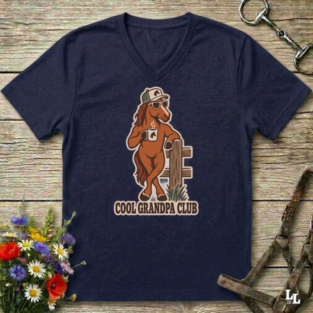 A navy blue v-neck t-shirt laid flat on a rustic wooden surface, featuring a cartoon graphic of a brown horse wearing sunglasses and a cap, holding a coffee mug next to a fence, with the text 'COOL GRANDPA CLUB' below. The shirt is styled with equestrian tack in the background and a small bouquet of flowers in the lower left corner.