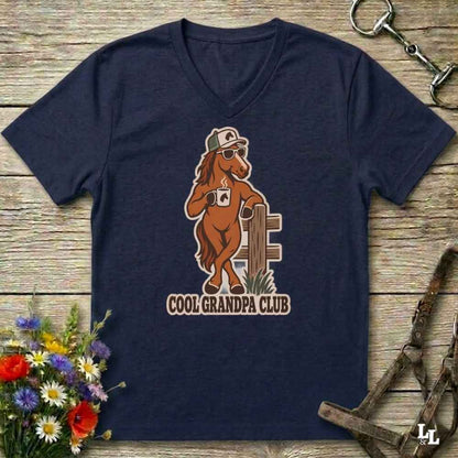 A navy blue v-neck t-shirt laid flat on a rustic wooden surface, featuring a cartoon graphic of a brown horse wearing sunglasses and a cap, holding a coffee mug next to a fence, with the text 'COOL GRANDPA CLUB' below. The shirt is styled with equestrian tack in the background and a small bouquet of flowers in the lower left corner.