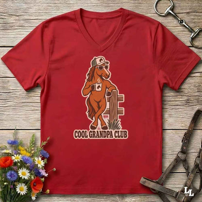 A red V-neck t-shirt laid flat on a rustic wooden surface, featuring a cartoon graphic of a cool brown horse wearing sunglasses and a cap, holding a coffee mug, with the text 'COOL GRANDPA CLUB' below. The shirt is styled with equestrian tack and a small flower arrangement.