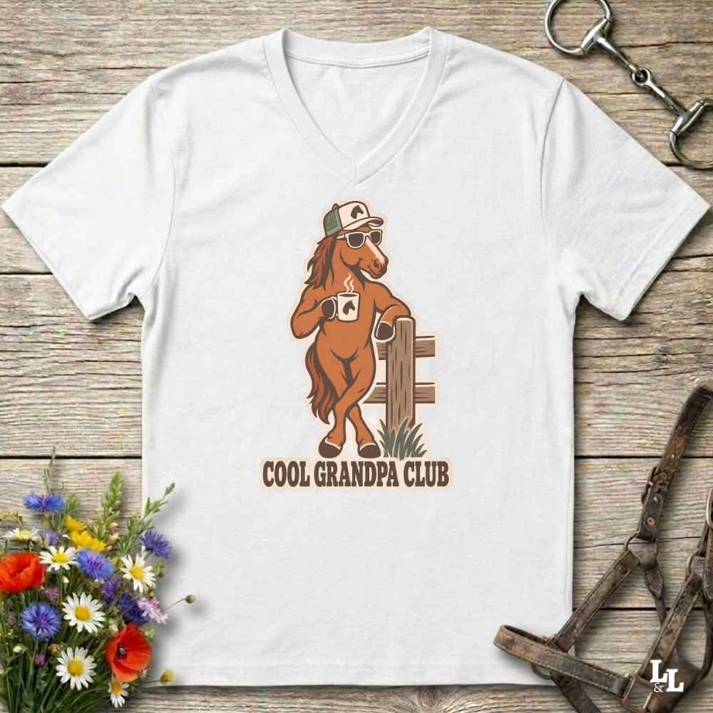 A white V-neck t-shirt laid flat on a rustic wooden surface, featuring a cartoon graphic of a brown horse wearing sunglasses and a hat, holding a coffee mug, with the text 'COOL GRANDPA CLUB' below. The shirt is styled with equestrian accessories (reins and a halter) and a small bouquet of wildflowers.