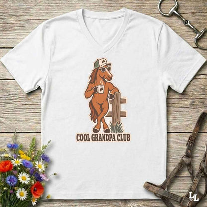 A white V-neck t-shirt laid flat on a rustic wooden surface, featuring a cartoon graphic of a brown horse wearing sunglasses and a hat, holding a coffee mug, with the text 'COOL GRANDPA CLUB' below. The shirt is styled with equestrian accessories (reins and a halter) and a small bouquet of wildflowers.