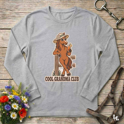 A heather gray long-sleeve t-shirt with a graphic of a brown horse wearing a cowboy hat and sunglasses, holding a drink, with the text 'COOL GRANDMA CLUB' underneath. The shirt is displayed in a flat-lay arrangement on a rustic wooden background with decorative flowers and leather tack.