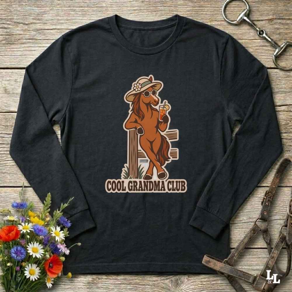 A black long-sleeve t-shirt with a graphic of a cool grandma horse and the text 'COOL GRANDMA CLUB', displayed on a rustic wooden background with equestrian accessories and flowers.