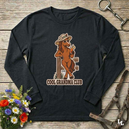 A black long-sleeve t-shirt with a graphic of a cool grandma horse and the text 'COOL GRANDMA CLUB', displayed on a rustic wooden background with equestrian accessories and flowers.