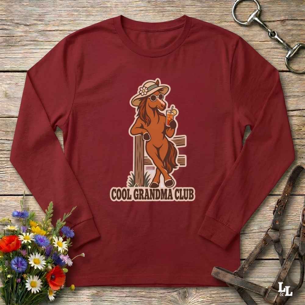 A long-sleeve crewneck t-shirt in maroon red, featuring a graphic of a cool horse wearing a hat and sunglasses while holding a drink, with the text 'COOL GRANDMA CLUB' underneath. The shirt is displayed flat on a rustic wooden background with equestrian and floral props.