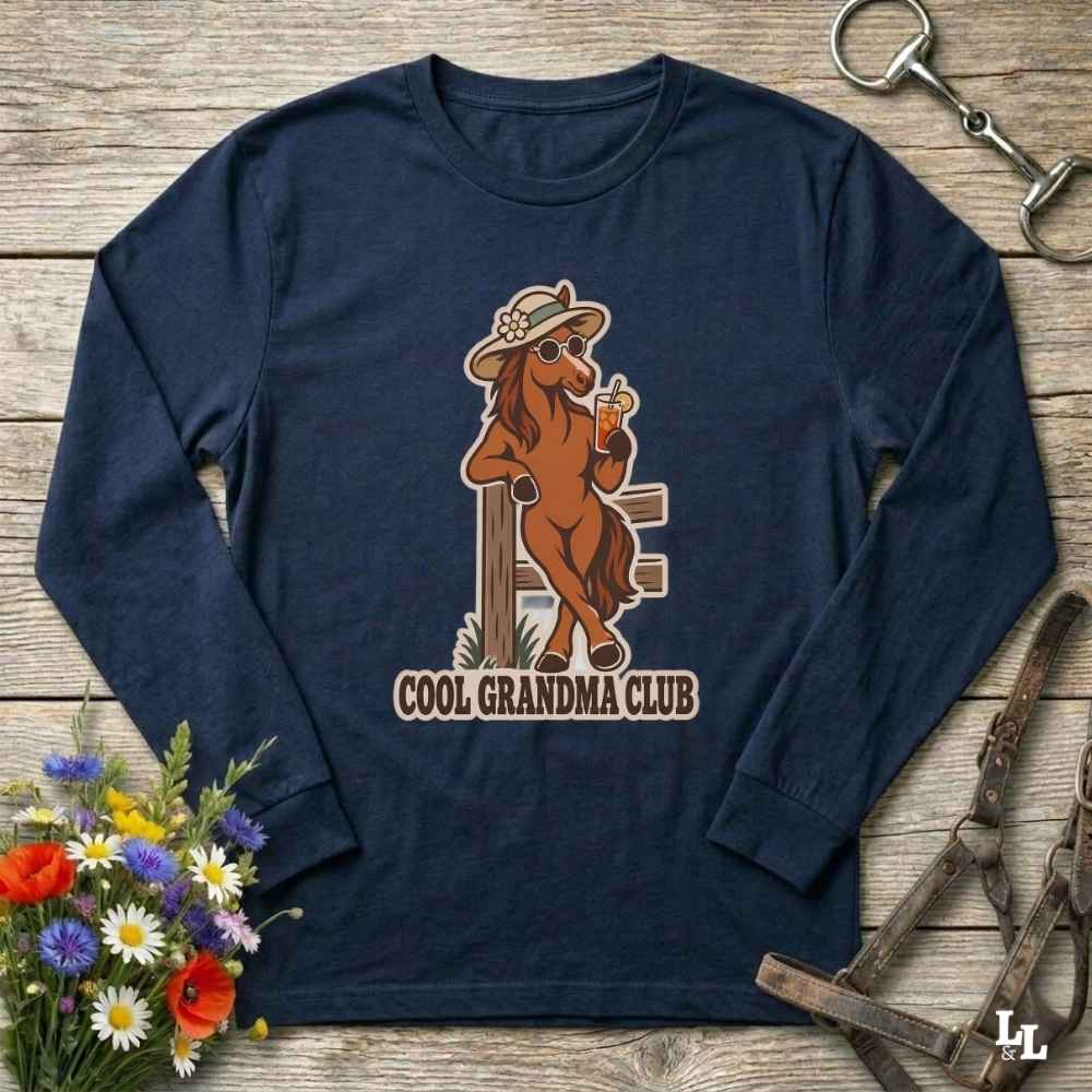 A navy blue long-sleeve t-shirt with a vintage-style graphic of a cool brown horse wearing a hat and sunglasses, holding a drink, with the text 'COOL GRANDMA CLUB' underneath. The shirt is displayed flat on a rustic wooden background with equestrian and floral props.