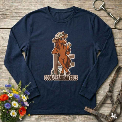 A navy blue long-sleeve t-shirt with a vintage-style graphic of a cool brown horse wearing a hat and sunglasses, holding a drink, with the text 'COOL GRANDMA CLUB' underneath. The shirt is displayed flat on a rustic wooden background with equestrian and floral props.