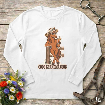A white long-sleeve crewneck t-shirt with a graphic of a cool brown horse wearing a hat and sunglasses, holding a drink, with the text 'COOL GRANDMA CLUB' underneath. The shirt is displayed flat on a rustic wooden background with equestrian accessories and flowers.
